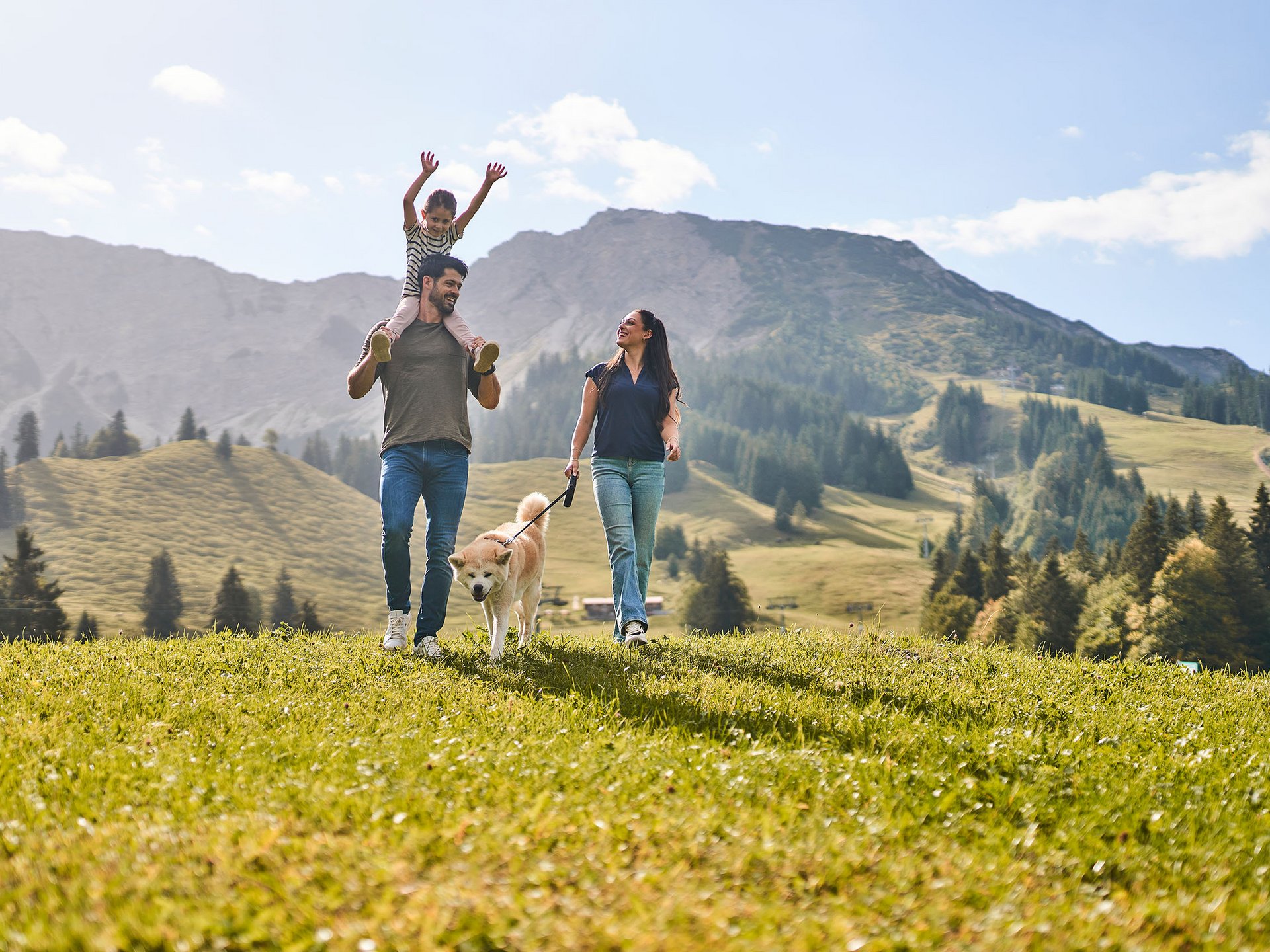 Familie mit Hund wandert durch eine alpine Wiese vor Bergpanorama